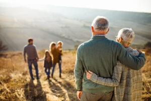 An elderly couple embraces while watching a young family walk through a sunlit field, symbolizing intergenerational health and well-being.